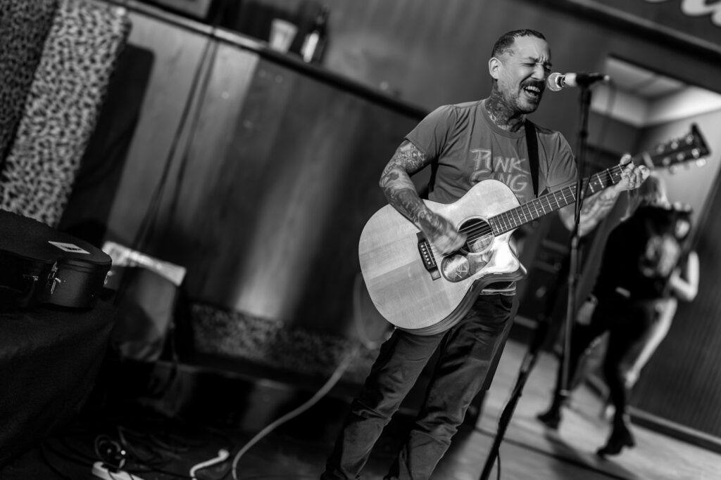 Photos show an assortment of nonbinary and male acoustic performers, some double exposed with bokeh on the audience. A small crowd fills the back room of a bar to watch them perform.